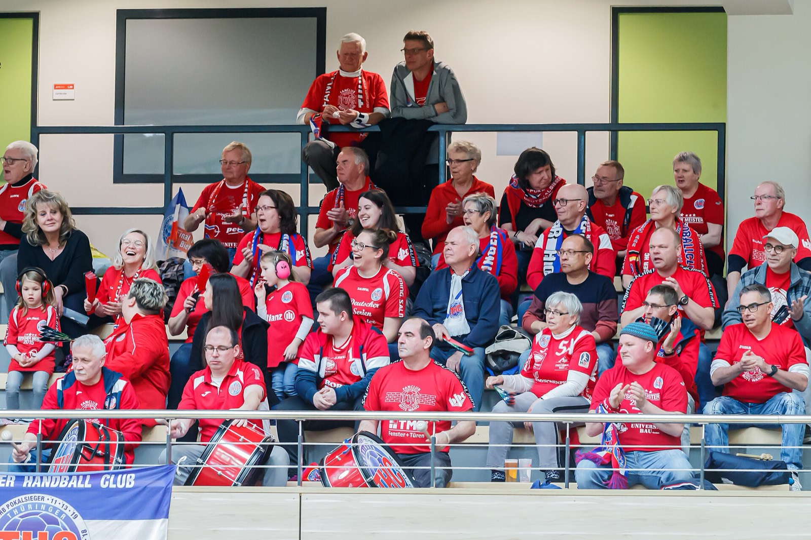 Zuschauer auf der Tribüne einer Handballhalle verfolgen ein Spiel, einige mit Trommeln und Fanschals